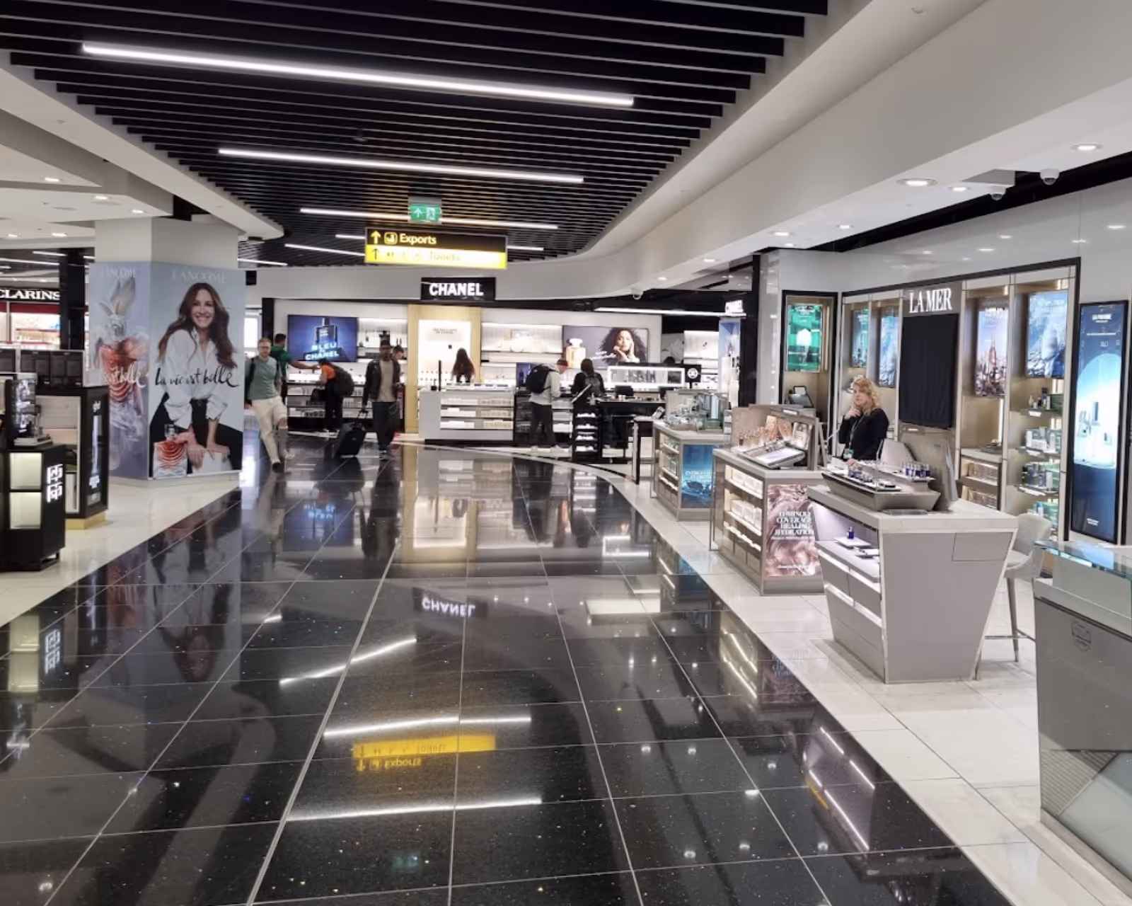 Travellers with luggage walk through a retail space in Gatwick Airport. Shops selling a variety of goods are on either side of a well-lit path lined with advertisements and promotions.